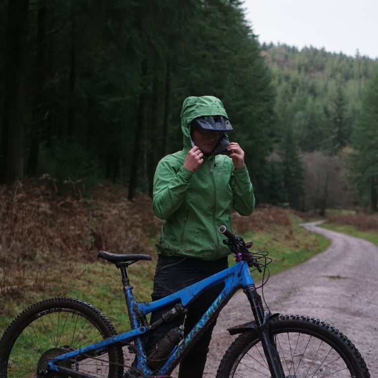 Rider in a rain-spotted Alpkit Balance jacket and hood fastens the collar beside a muddy blue bike on a gravel forest road.