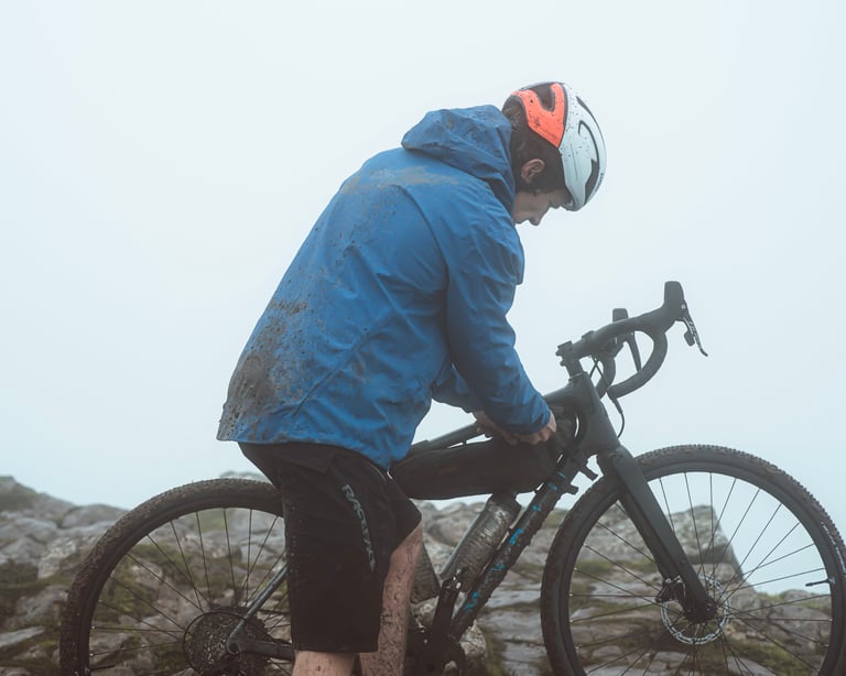Mud-splashed dhb Trail waterproof jacket on a rider with a gravel bike on a misty, rocky hilltop.