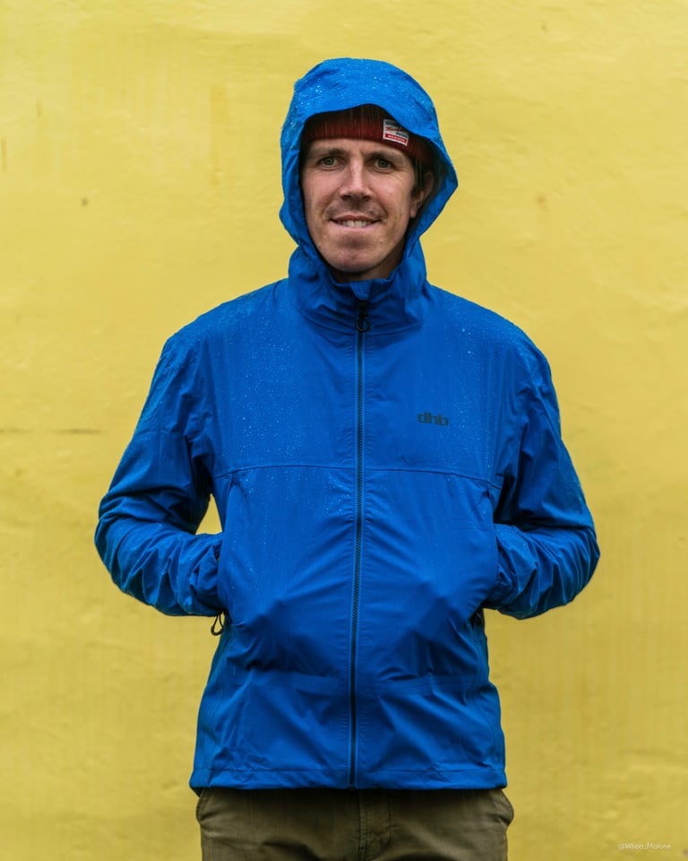 Rider wearing the blue dhb Trail waterproof jacket with the hood up and rain beading on the fabric.