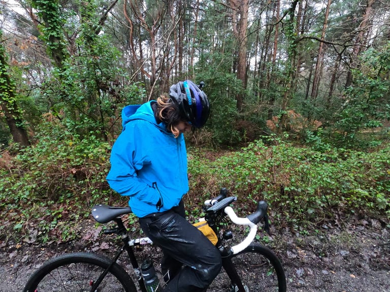 Rider stands astride a gravel bike in woodland with both hands tucked into the Madison DTE jacket's front pockets.