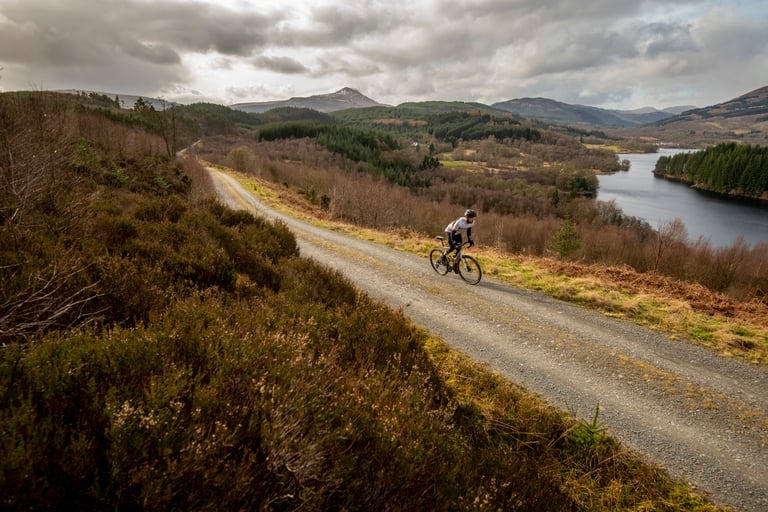 Rider on a winding gravel forest road above a loch near Aberfoyle, with snow-dusted mountains beyond.