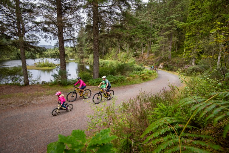 Family with a young child ride a forest gravel road beside a loch on a Gravelfoyle waymarked route near Aberfoyle.