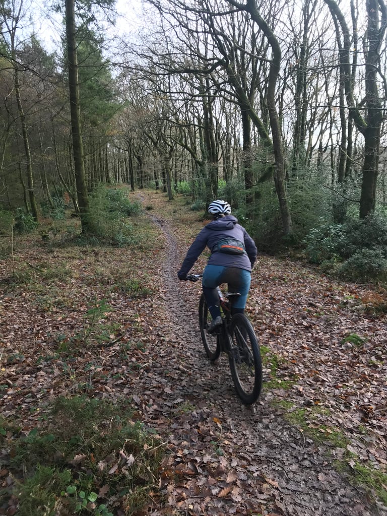 Rider in a Findra Stroma jacket and a small hip pack riding away along a leaf-covered woodland singletrack.