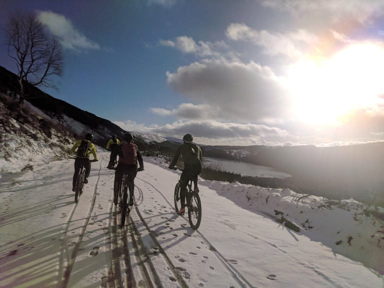 Four riders on an off-road Festive 500 follow a snowy hillside track above a lake in low winter sun.