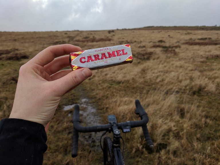 Hand holds a Tunnock's Caramel wafer over drop handlebars on open moorland during an off-road Festive 500 ride.