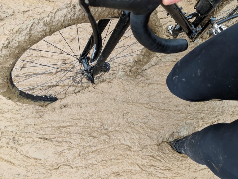 View down over a bike's front wheel and rider's leg stuck in deep, sloppy mud on an off-road winter route.