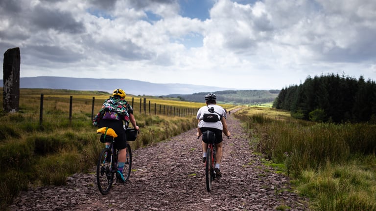 Two bikepackers ride a stony track past a standing stone on Sarn Helen, a bikepacking route through Wales.