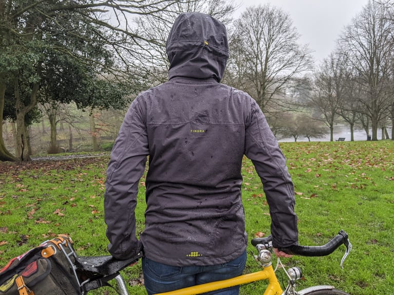 Back of the Findra Stroma jacket with hood up, rain beading on the grey fabric, the wearer holding a yellow bike.