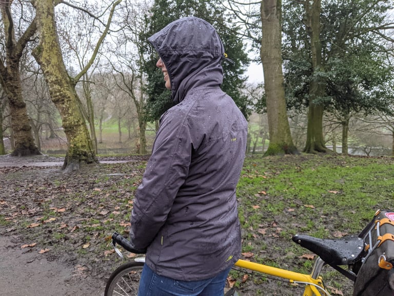 Side view of the Findra Stroma jacket in nine iron grey, hood up, beside a yellow bike in a rainy park.