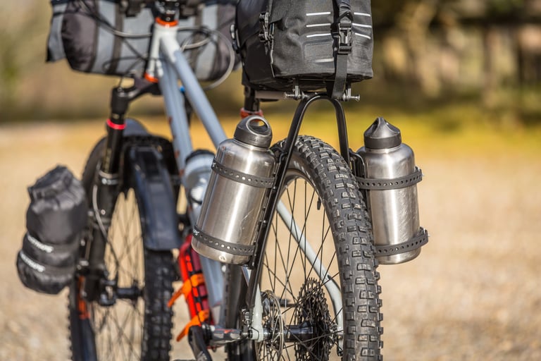 Two steel water bottles strapped into Tailfin Cargo Cages on the legs of a loaded bikepacking bike's rear rack.