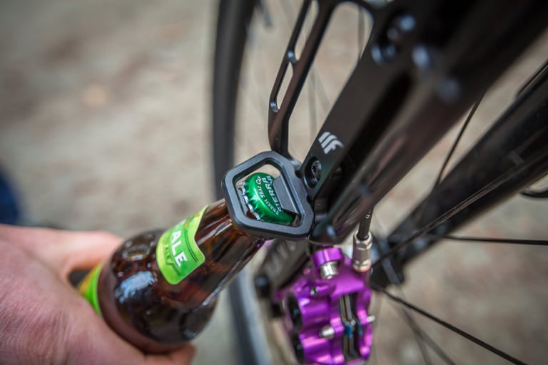Tailfin Cargo Cage Load Chip doubling as a bottle opener on a fork, popping the cap off a bottle of ale.