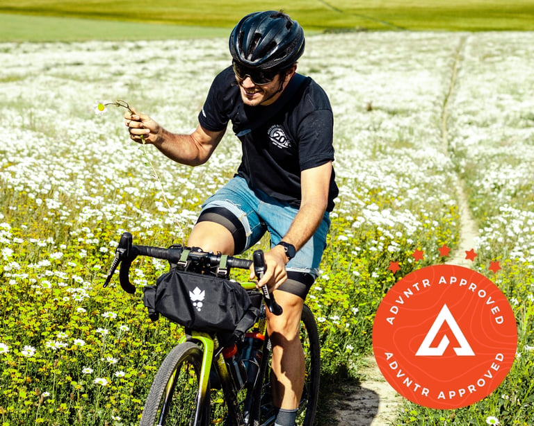 Rider in Ripton V2 Jorts holds a daisy while riding a gravel bike with a handlebar bag through a daisy meadow.