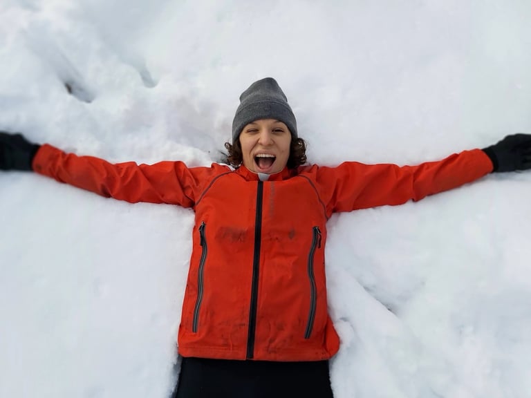 Rider in the red Showers Pass Refuge jacket makes a snow angel, arms outstretched in fresh snow.