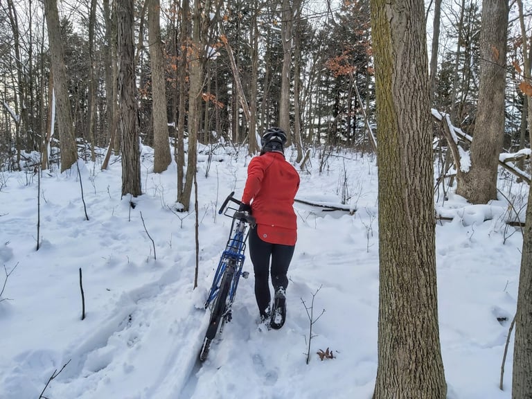 Rider in a red jacket and Shimano MW7 winter boots pushes a blue bike through snowy woodland.