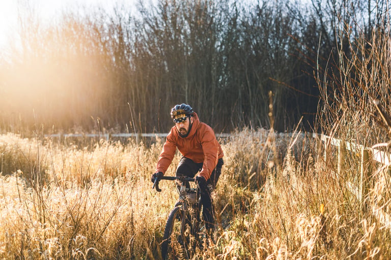 Rider in the relaxed-fit Copilot jacket on a gravel bike through tall golden grass in low winter sun.