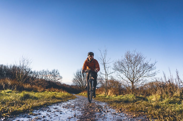 Rider in the Copilot waterproof jacket riding towards the camera on a puddled gravel track under a clear blue sky.