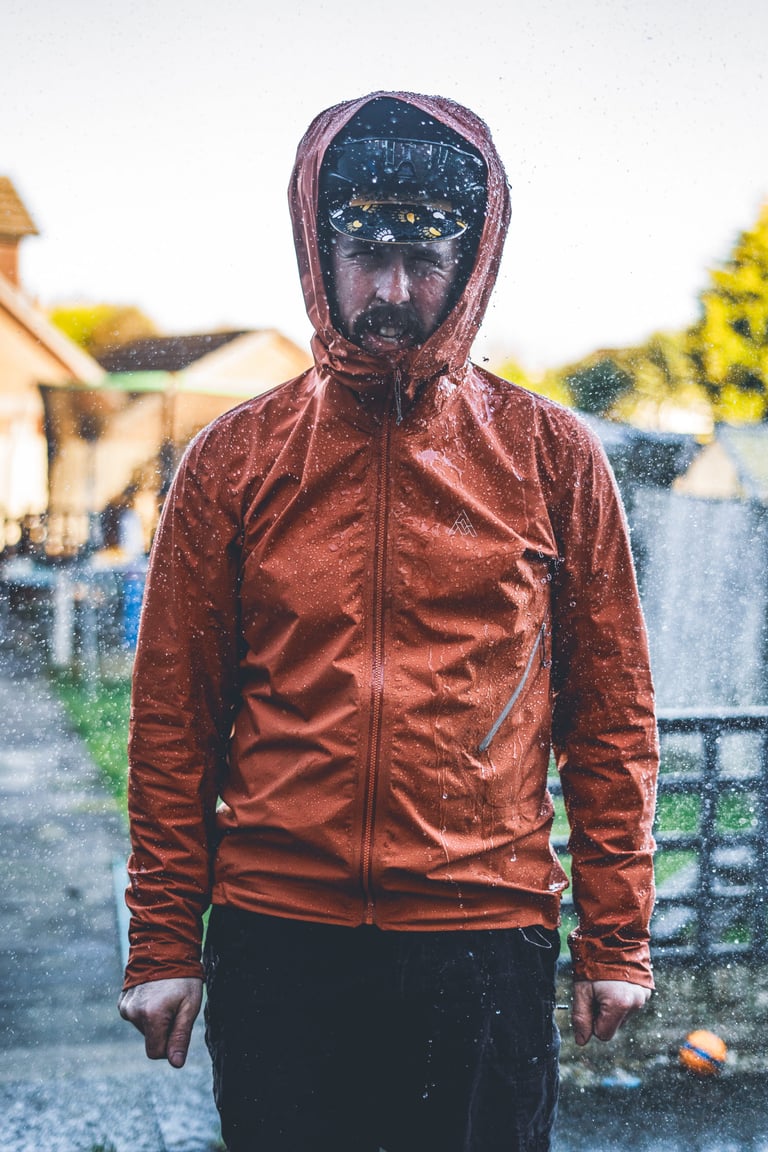 7mesh Copilot jacket with its hood up, soaked by a garden hose, with water running off the orange shell.