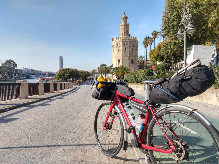Red Norco Search loaded with bikepacking bags on the riverside in Seville, with a historic stone tower behind.