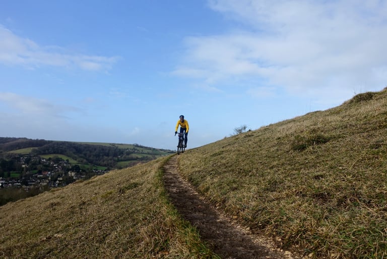 Rider in a yellow Velocio jacket descends a narrow grassy singletrack on a Cotswold hillside above a village.
