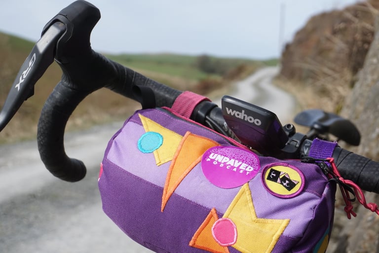 Wahoo ELEMNT ROAM on its out-front mount beside a purple, patched handlebar bag on a gravel lane.