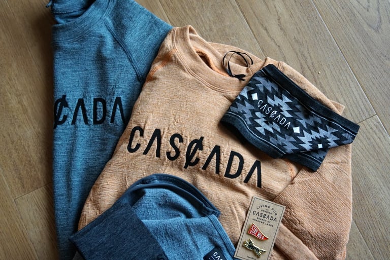 Cascada merino baselayers in grey-blue and peach with a patterned headband and pin badges on a wooden floor.