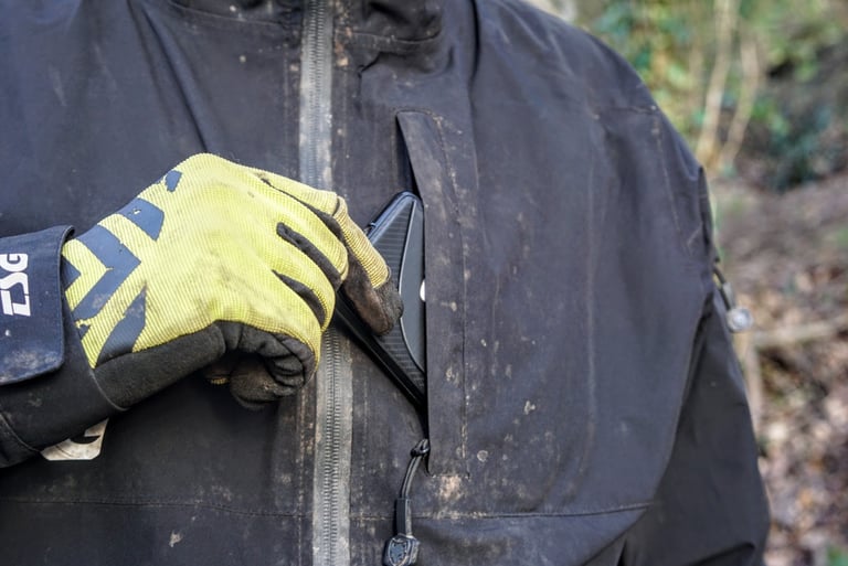 Gloved hand slides a smartphone into the zipped chest pocket of the muddy TSG Drop Rain Jacket.