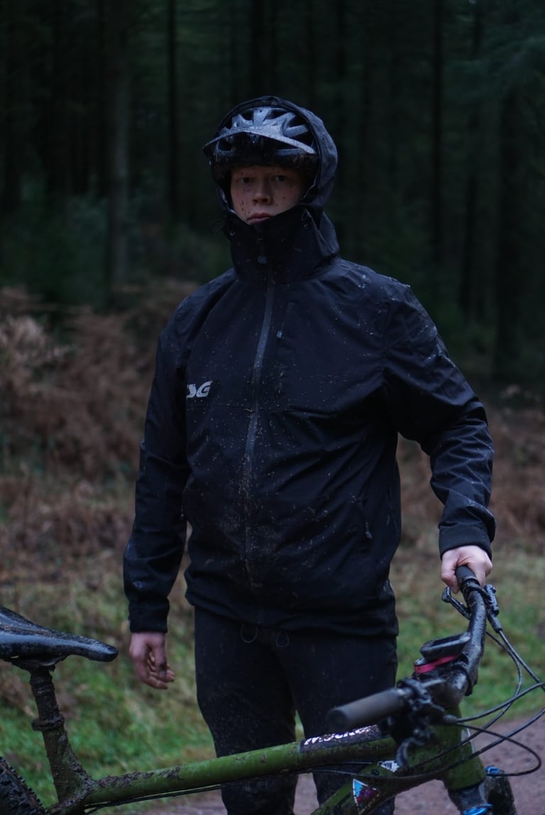 Rider wearing the size L TSG Drop Rain Jacket, hood up over a helmet, beside a muddy mountain bike in the forest.