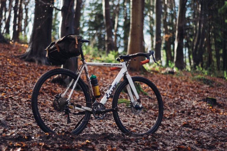 White Cube bike labelled 'Gravel Amateur', with a saddle bag and bottle, parked among fallen leaves in woodland.