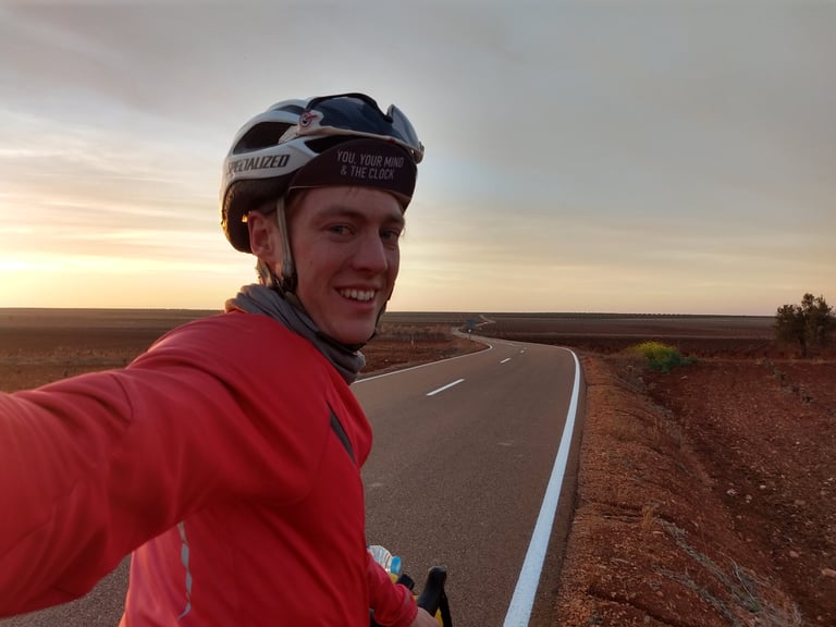 Ceri Middleton takes a smiling selfie in a red jacket on an empty Spanish road through red farmland at sunset.