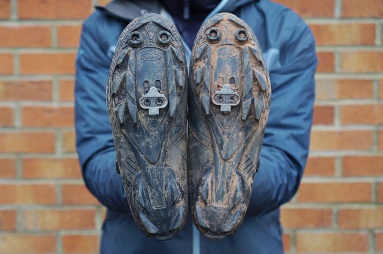 Muddy soles of Northwave Magma XC Core shoes held up to the camera, showing the stiff tread and cleats.