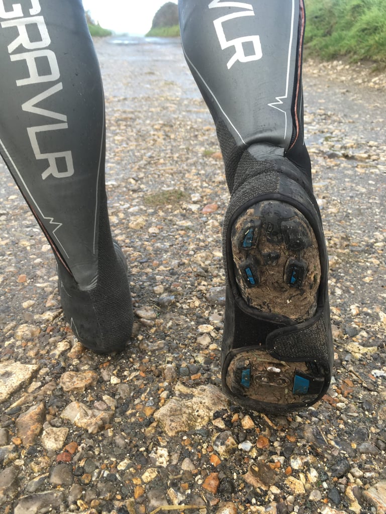 Muddy sole of a SPATZ GravlR overshoe lifted to show the cleat and the underfoot velcro strap on a wet track.
