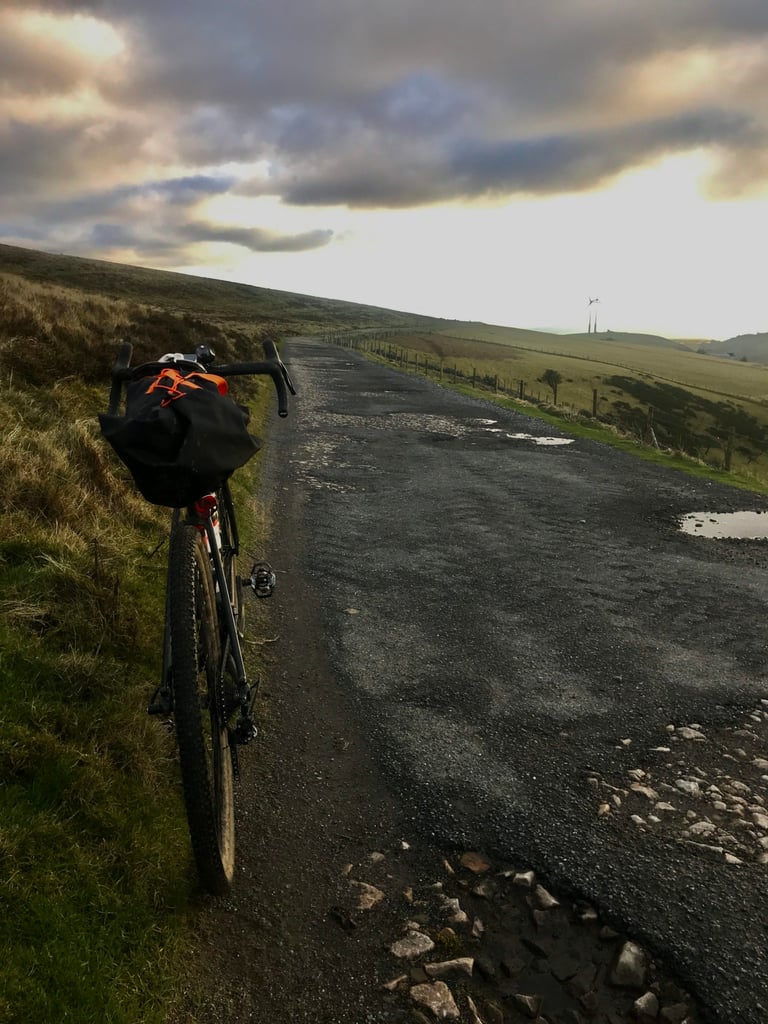 Gravel bike with a handlebar bag on a broken tarmac ridge road with wind turbines ahead, South Wales valleys.