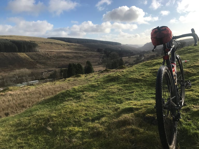 Gravel bike with a red handlebar bag on a grassy hilltop above forested South Wales valleys.