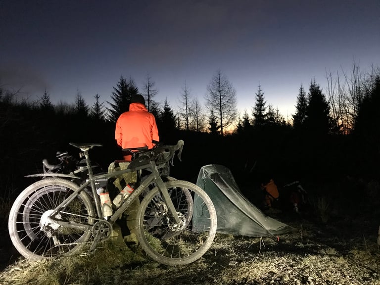 Rider in an orange jacket beside a gravel bike and a bivvy tent at dusk, on the Llanwonno family campout.
