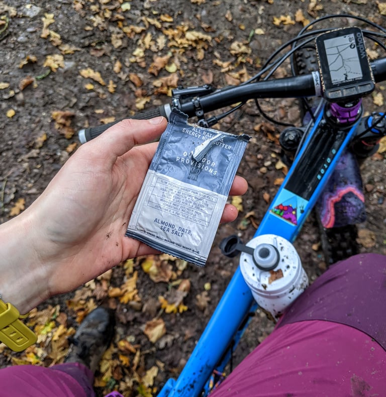 Rider holds a torn-open Outdoor Provisions almond, date and sea salt sachet over a muddy blue mountain bike.