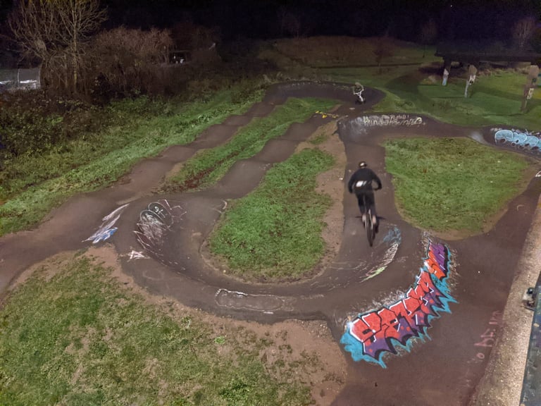 A rider rounds a banked, graffiti-covered corner of a pump track at night, seen from above.