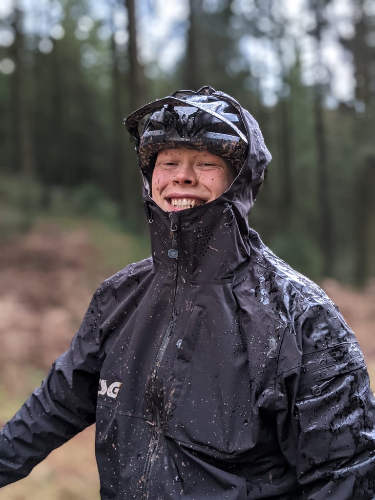 Smiling rider in a rain-soaked, mud-spattered TSG Drop Rain Jacket with hood up over a helmet in a wet forest.