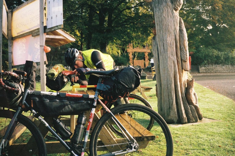 Rider in a hi-vis gilet leans over a loaded touring bike at a picnic bench beside a carved wooden tree trunk.