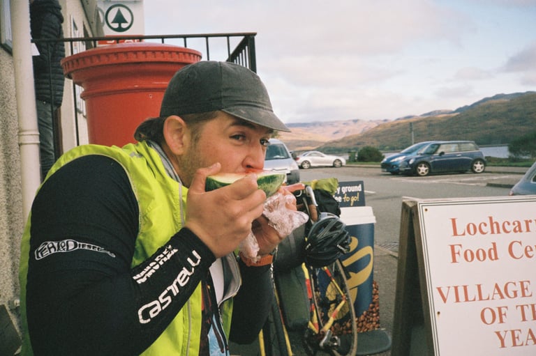 Lewis eats a slice of watermelon outside a shop in Lochcarron, his touring bike propped behind him.