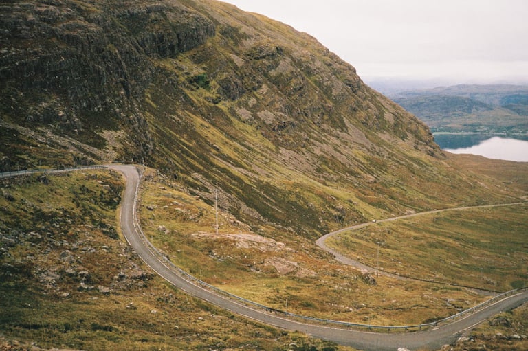Narrow road twists in hairpin bends up a steep hillside on the Bealach na Bà, with a loch far below.