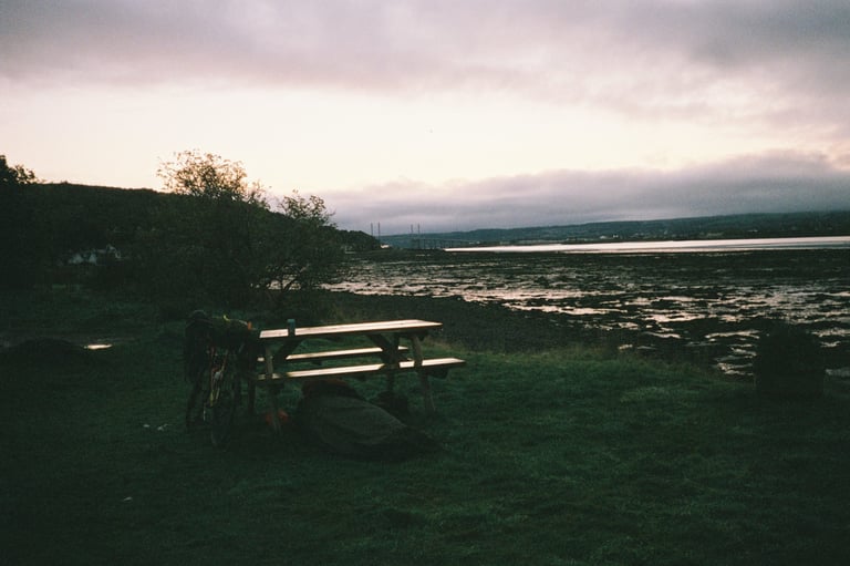 Picnic bench and a loaded bike on the shore at North Kessock near Inverness at dusk, with a bridge in the distance.