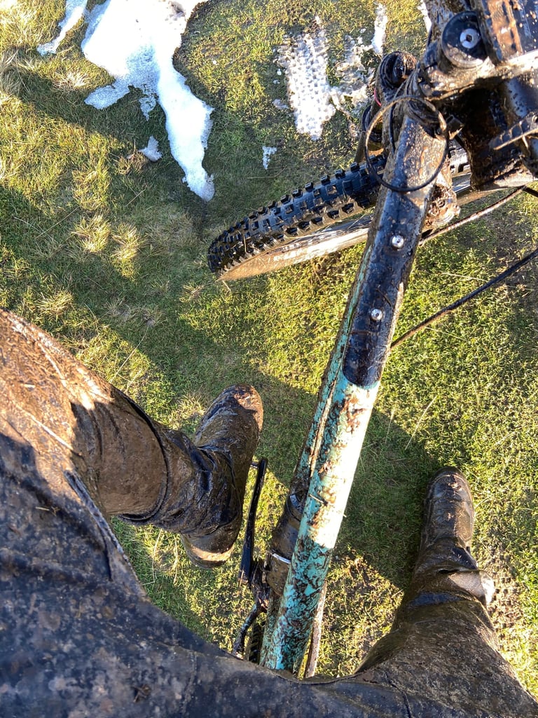 Looking down from the saddle at the Cale AL's top tube and its two bag-mount bolts, with muddy legs and patchy snow.