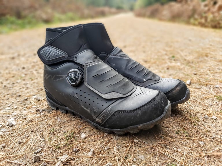 Pair of black Shimano MW7 winter boots on a gravel track, with BOA dial, perforated toe box and tall cuff.