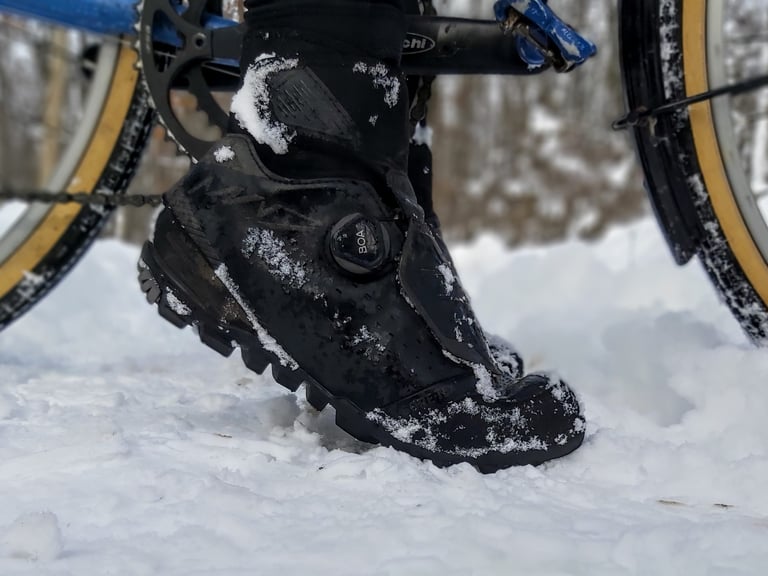 Rider's Shimano MW7 winter shoe clipped into the pedal of a blue bike, with snow on the boot and tyres.
