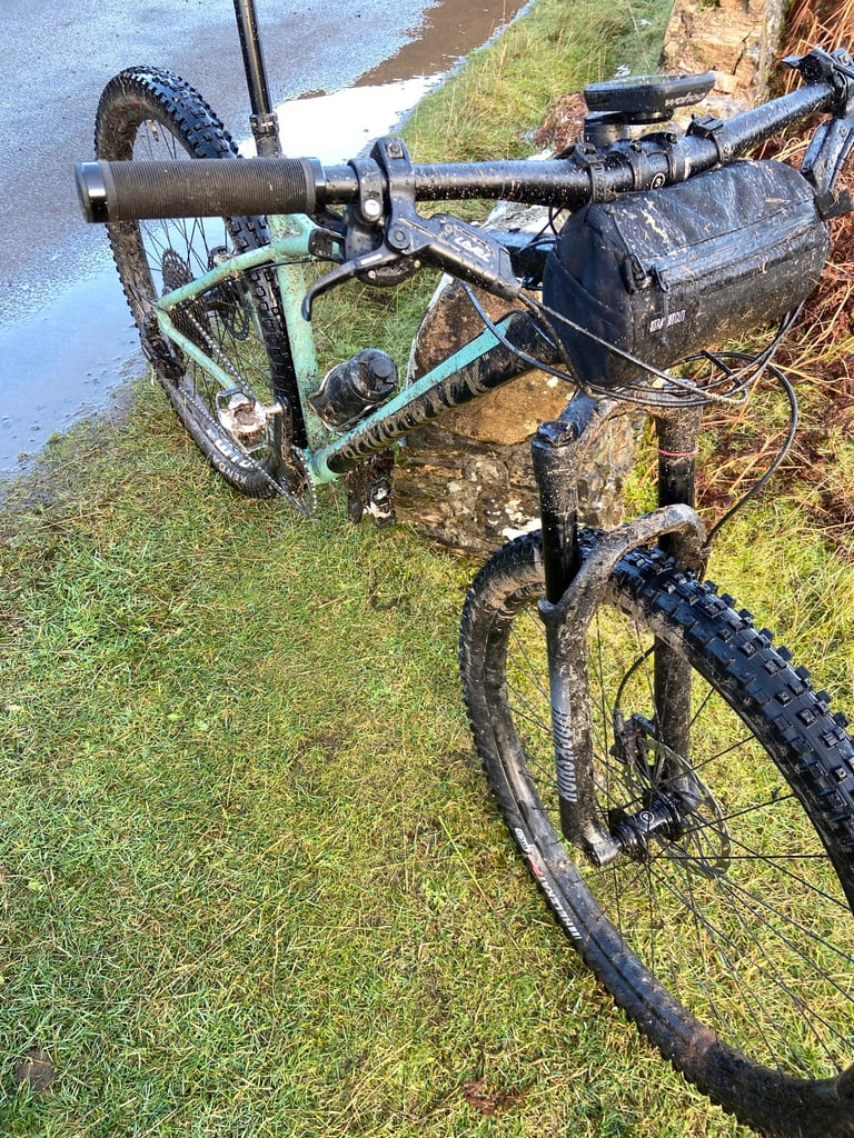 Mud-spattered Cale AL cockpit with wide bars, grips and a handlebar bag beside a stone wall.