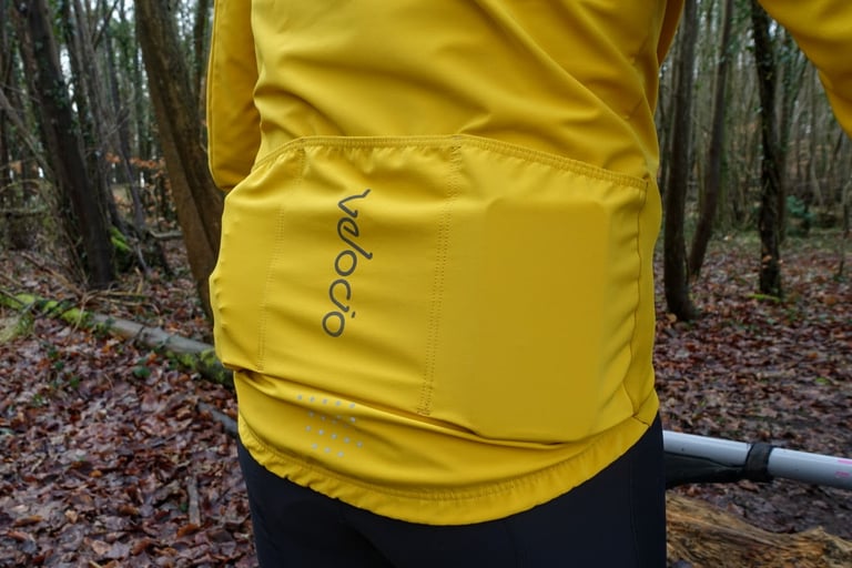 Rear of the yellow Velocio Signature soft shell jacket, with three rear pockets, Velocio logo and reflective hem dots.