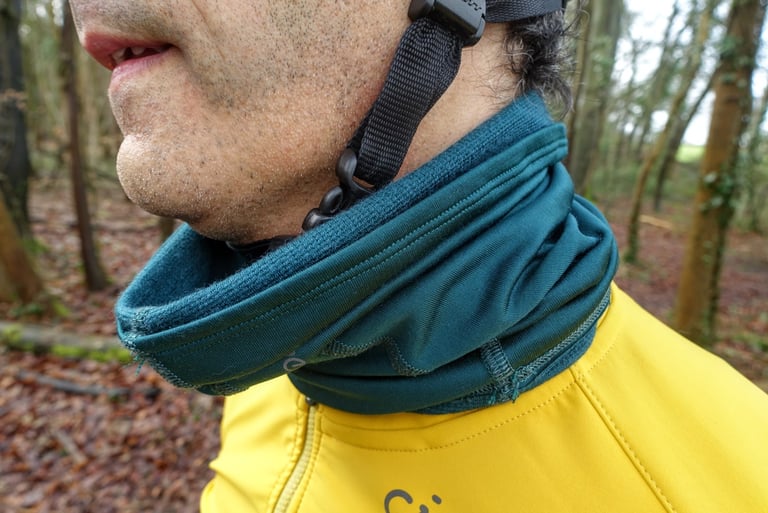 Dark green Velocio Merino 210 winter collar worn as a neck gaiter over a yellow soft shell, with a helmet strap beside it.