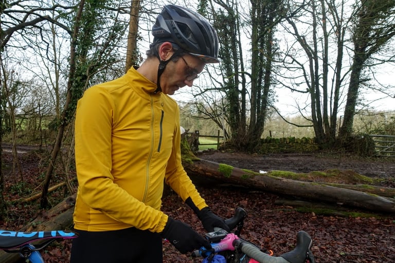 Rider in a yellow Velocio Signature soft shell jacket and black gloves checks the bike computer on a gravel bike.