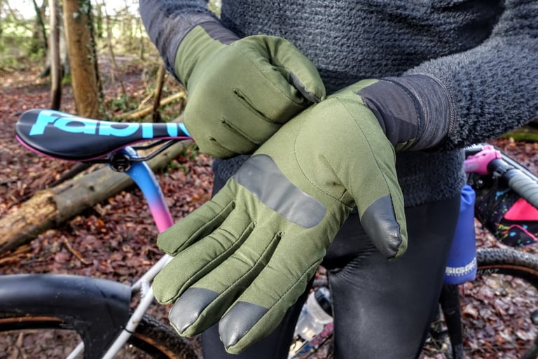 Pair of olive green Velocio Alpha gloves with grey palm patches and fingertips, being pulled on beside a gravel bike.