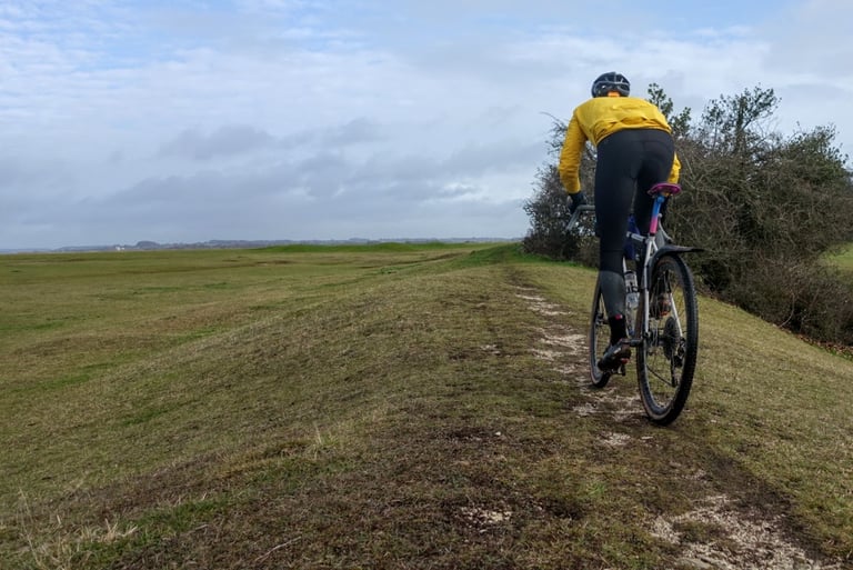 Rider in a yellow Velocio jacket and Zero bib tights rides a gravel bike along a grassy ridge track.